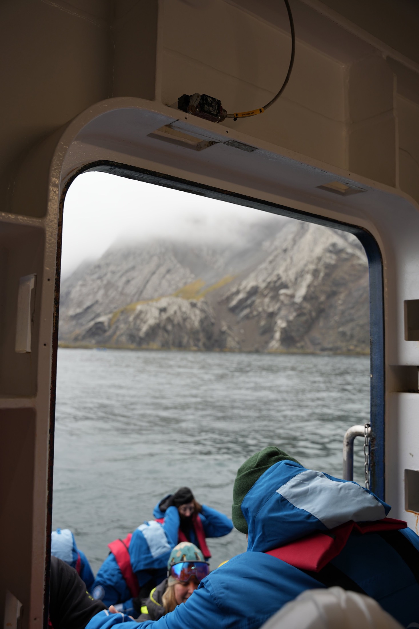 Through a ship's porthole: travellers in red and blue expedition jackets boarding a Zodiac below misty cliffs in South Georgia