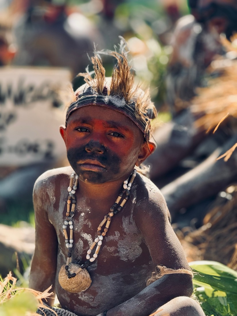 A small boy blackened with charcoal, feathers in his headband and shells on his chest, sits among dancers in Papua New Guinea