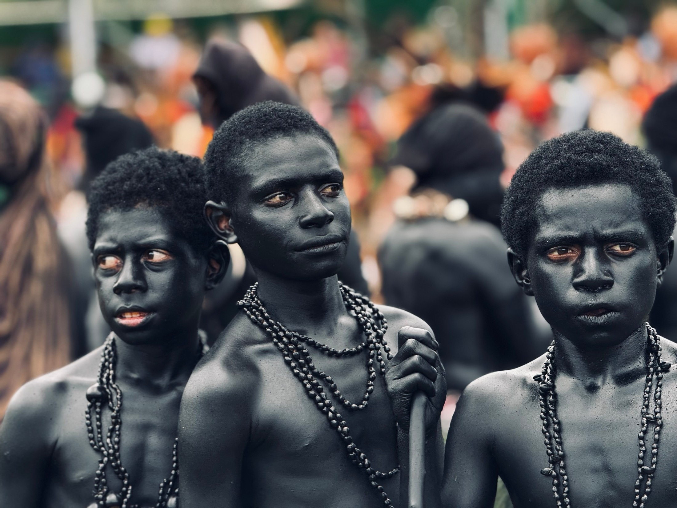 Three boys blackened with charcoal and wearing seed necklaces stand together in a festival crowd in Papua New Guinea