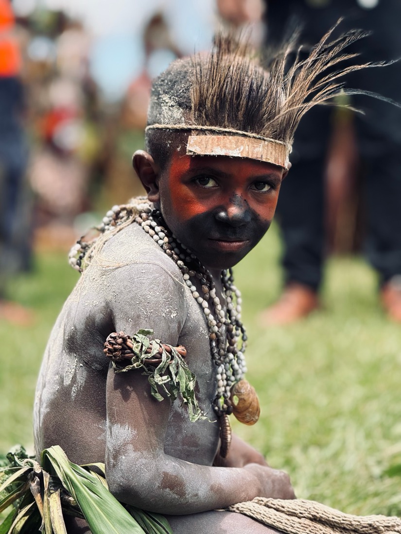 A boy painted in grey clay with a feathered headband and shell necklaces looks back over his shoulder, Papua New Guinea