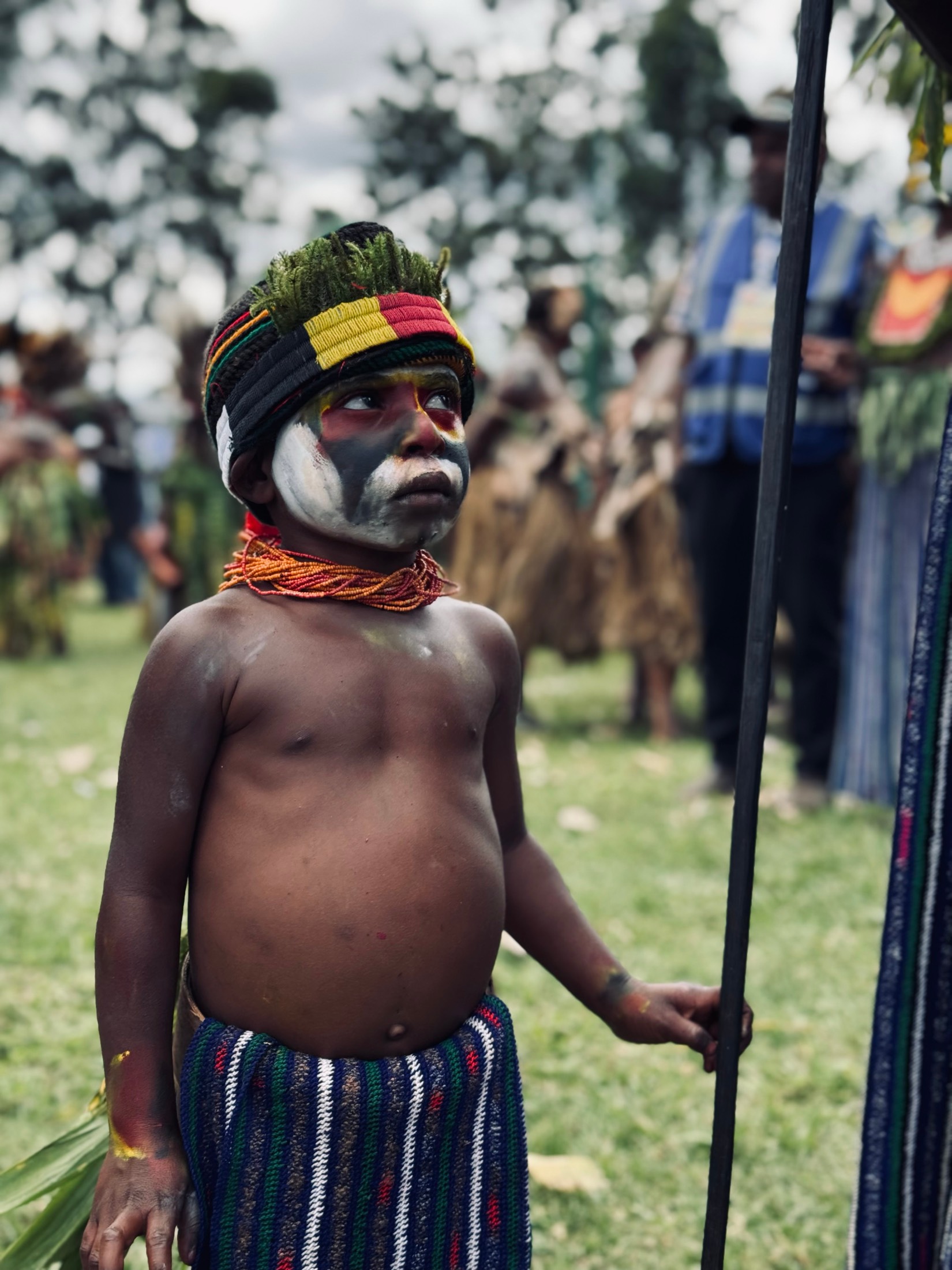 A young boy in face paint and a striped sarong stands holding a tall spear at a Highlands sing-sing, Papua New Guinea