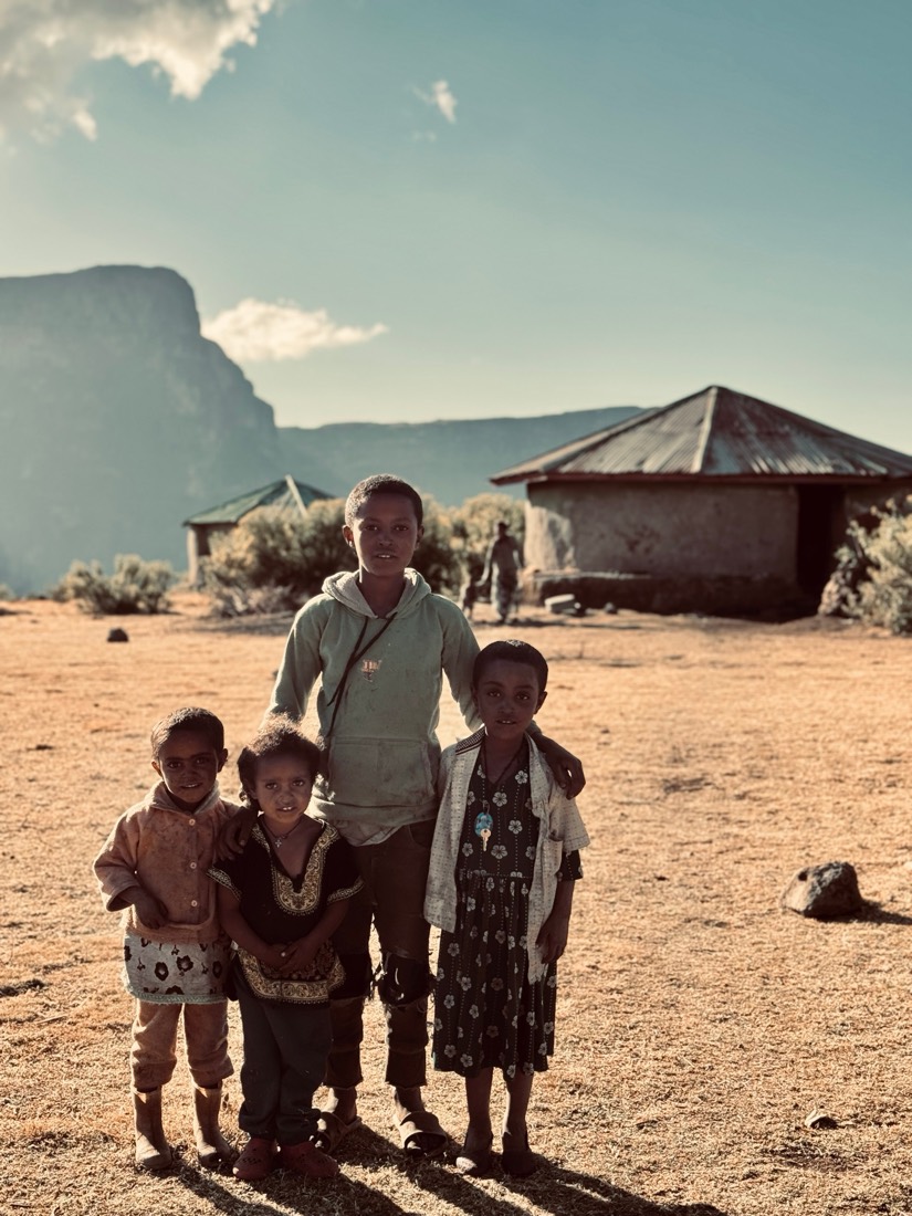 A young man with three small children outside round stone huts, escarpment cliffs behind, in the Simien Mountains of Ethiopia