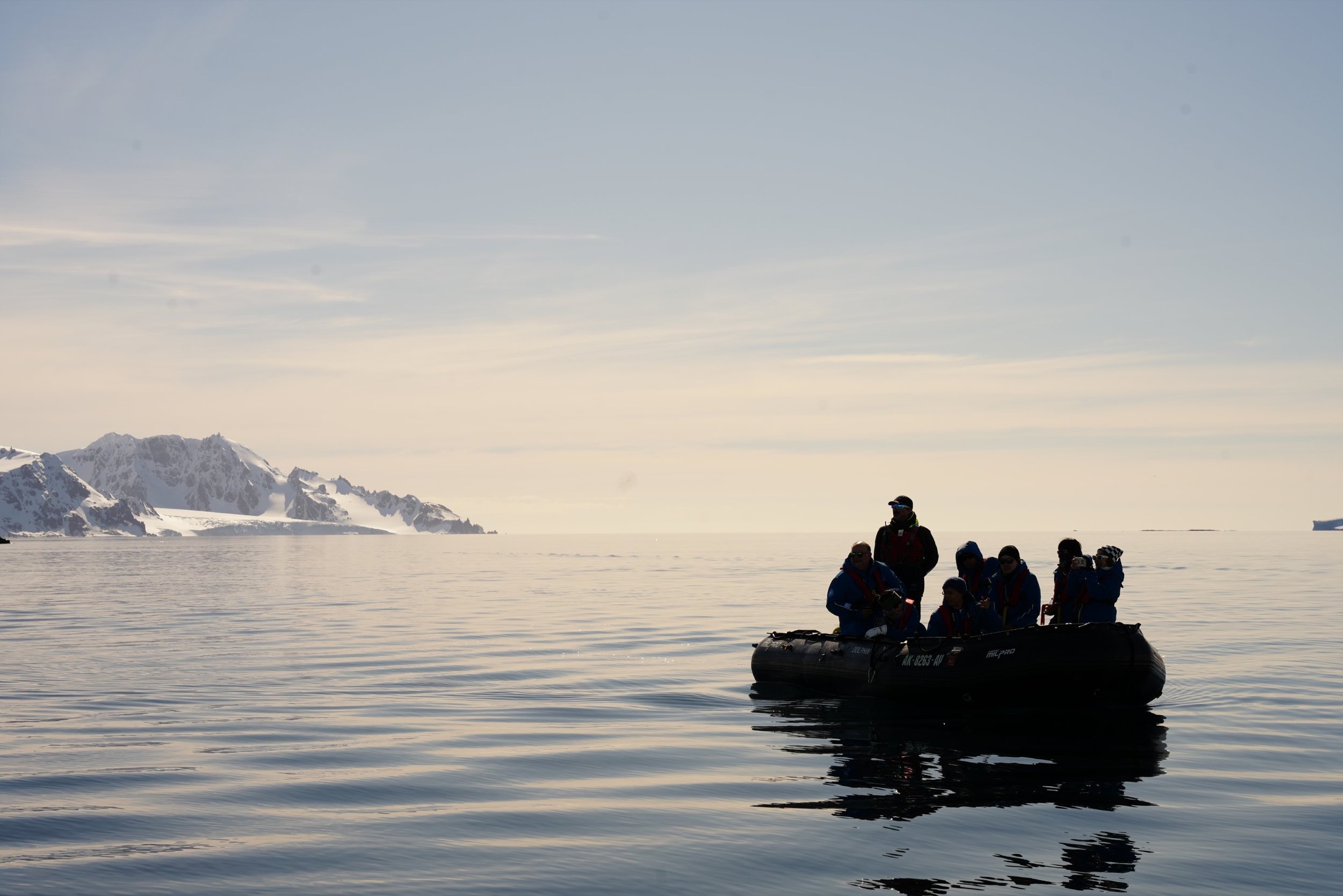 Zodiac full of travellers silhouetted on calm water, snowy Antarctic coastline behind