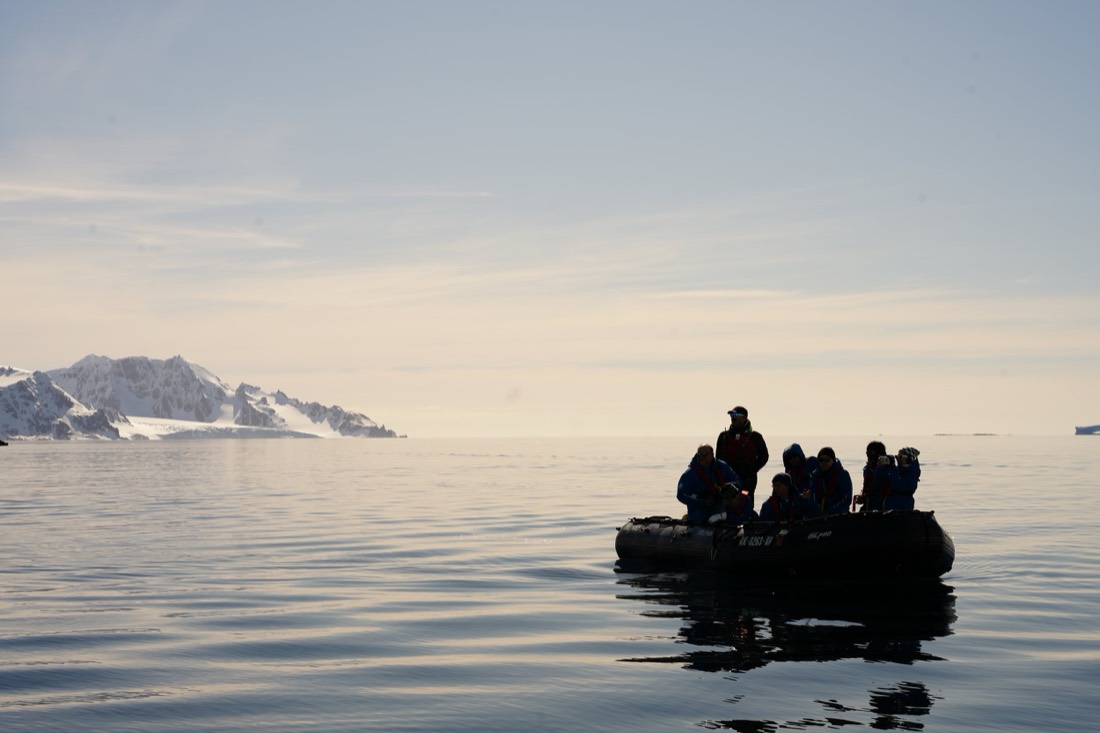 Zodiac full of travellers silhouetted on calm water, snowy Antarctic coastline behind