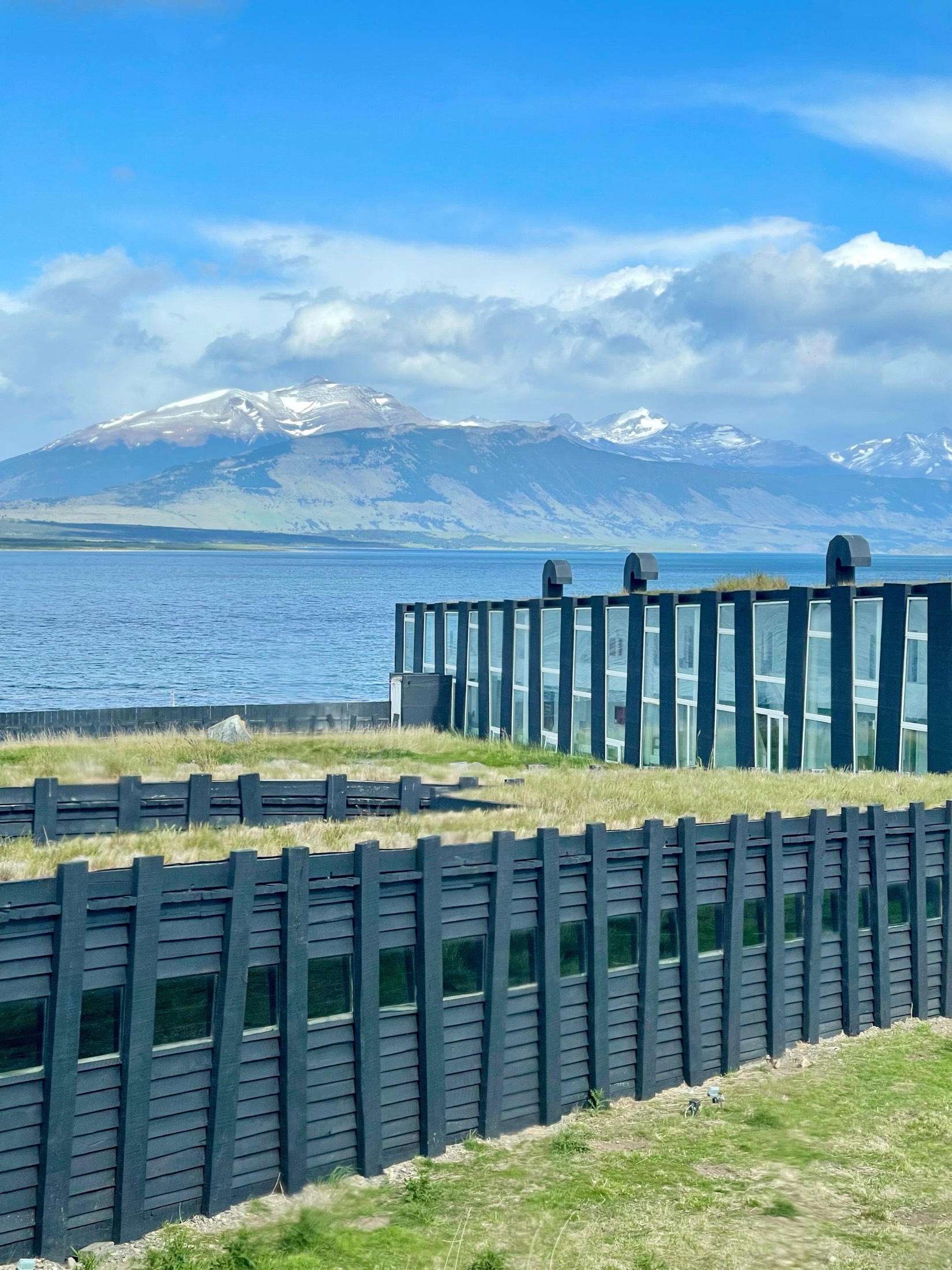 Dark timber slats and a grass roof step down to a blue lake, snow-capped peaks beyond, Chilean Patagonia