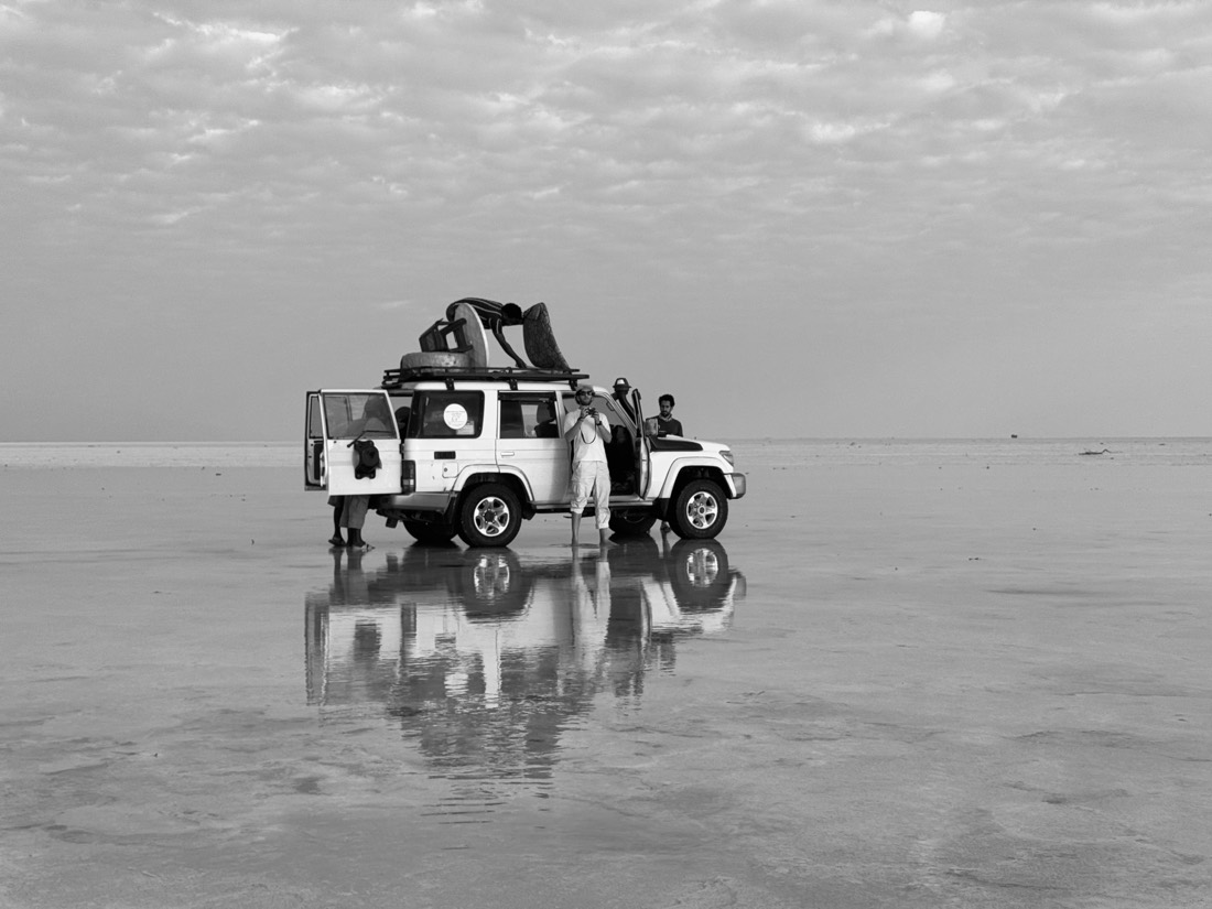 A loaded four-by-four and its reflection stand on a flooded salt flat under a grey sky, in black and white