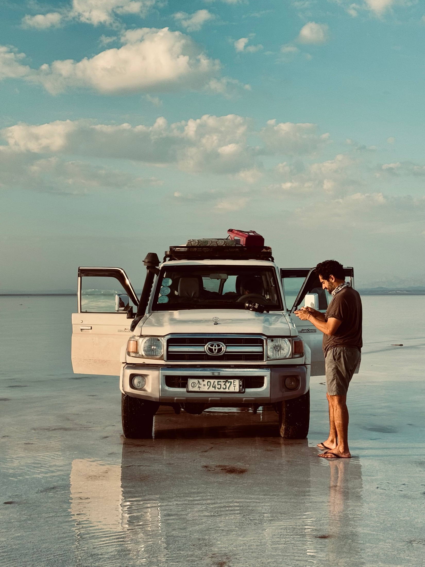 A man checks his phone beside a Land Cruiser with its doors open on a shining wet salt flat in the Danakil Depression