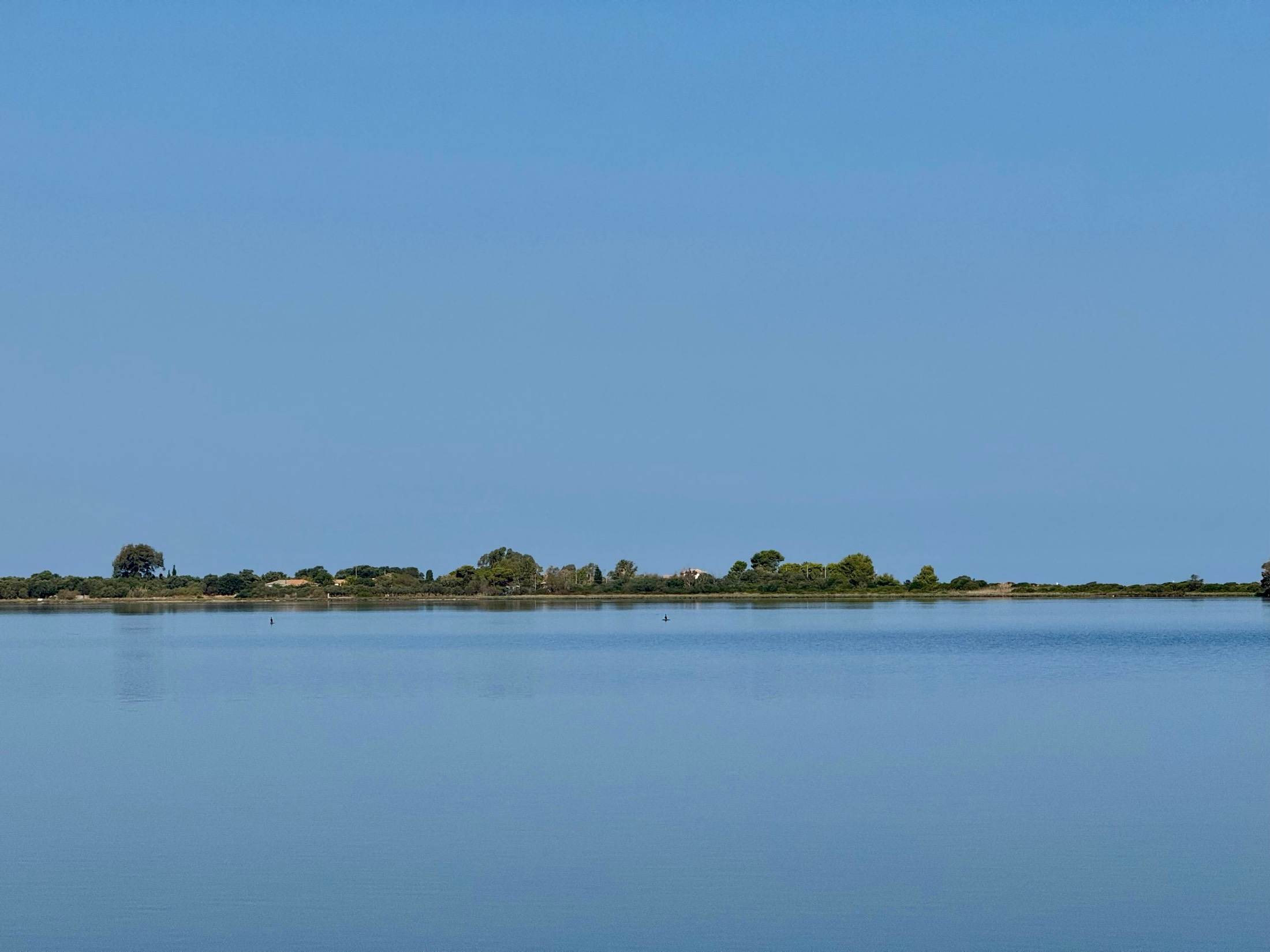 A low green shoreline floats between a still lagoon and a pale blue sky, Greece