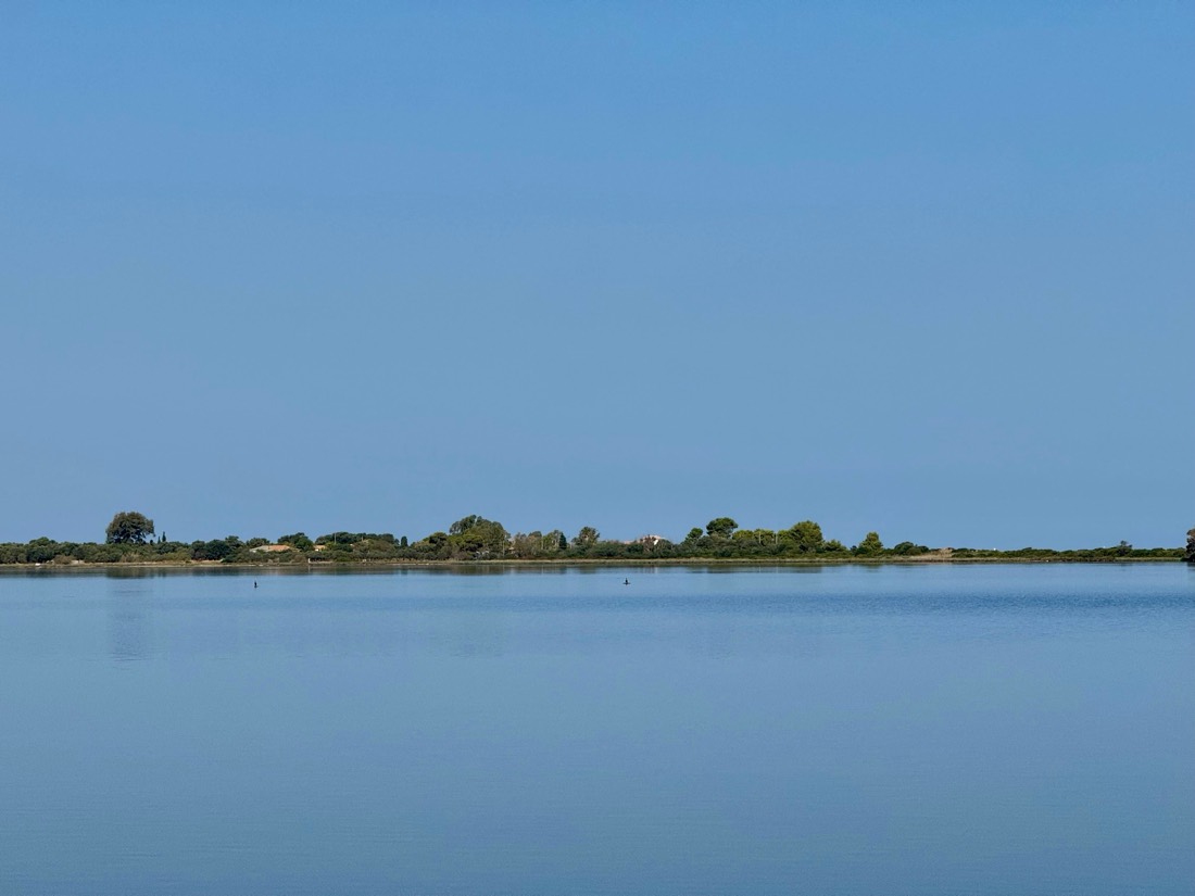 A low green shoreline floats between a still lagoon and a pale blue sky, Greece
