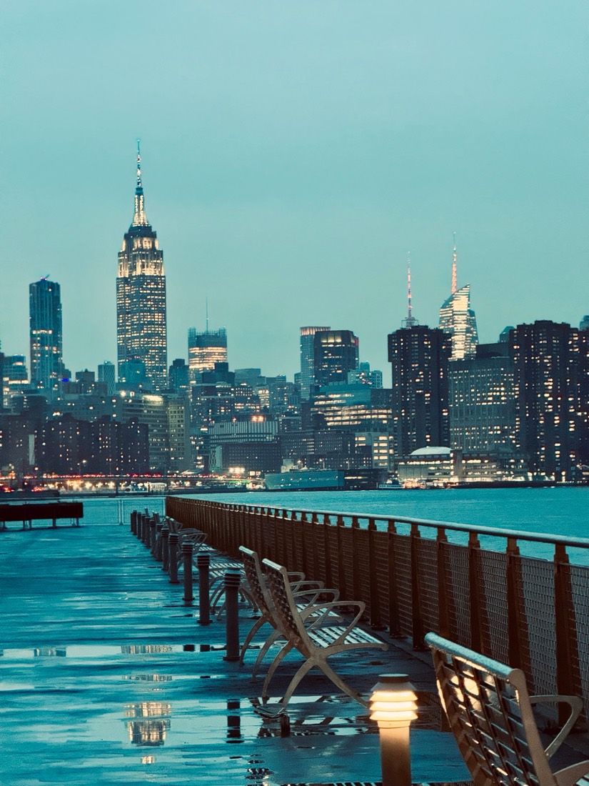 A wet wooden pier leads toward the Manhattan skyline and the Empire State Building at dusk