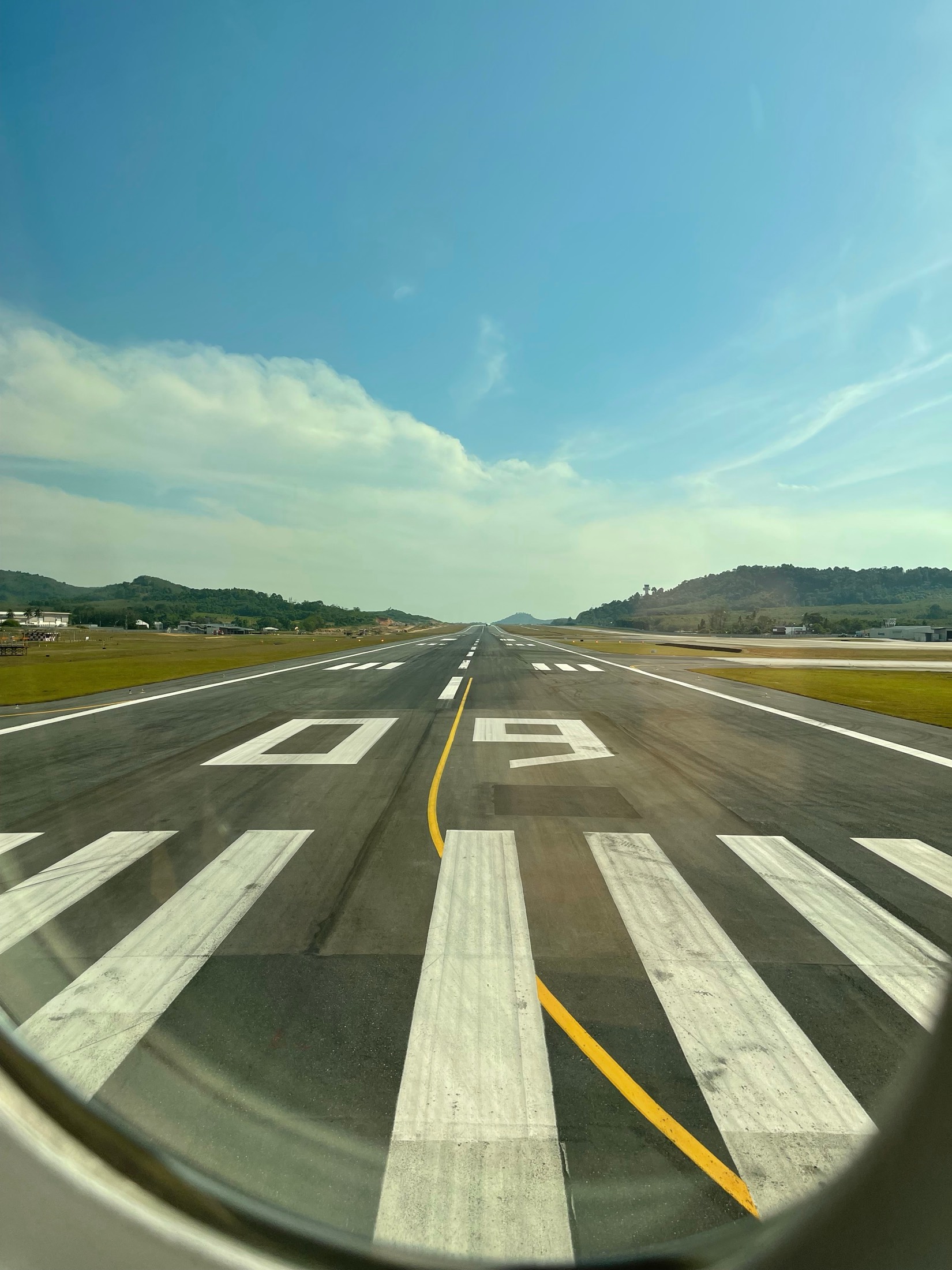 The painted threshold of a runway and green hills ahead, seen from the cockpit side of a plane, Thailand