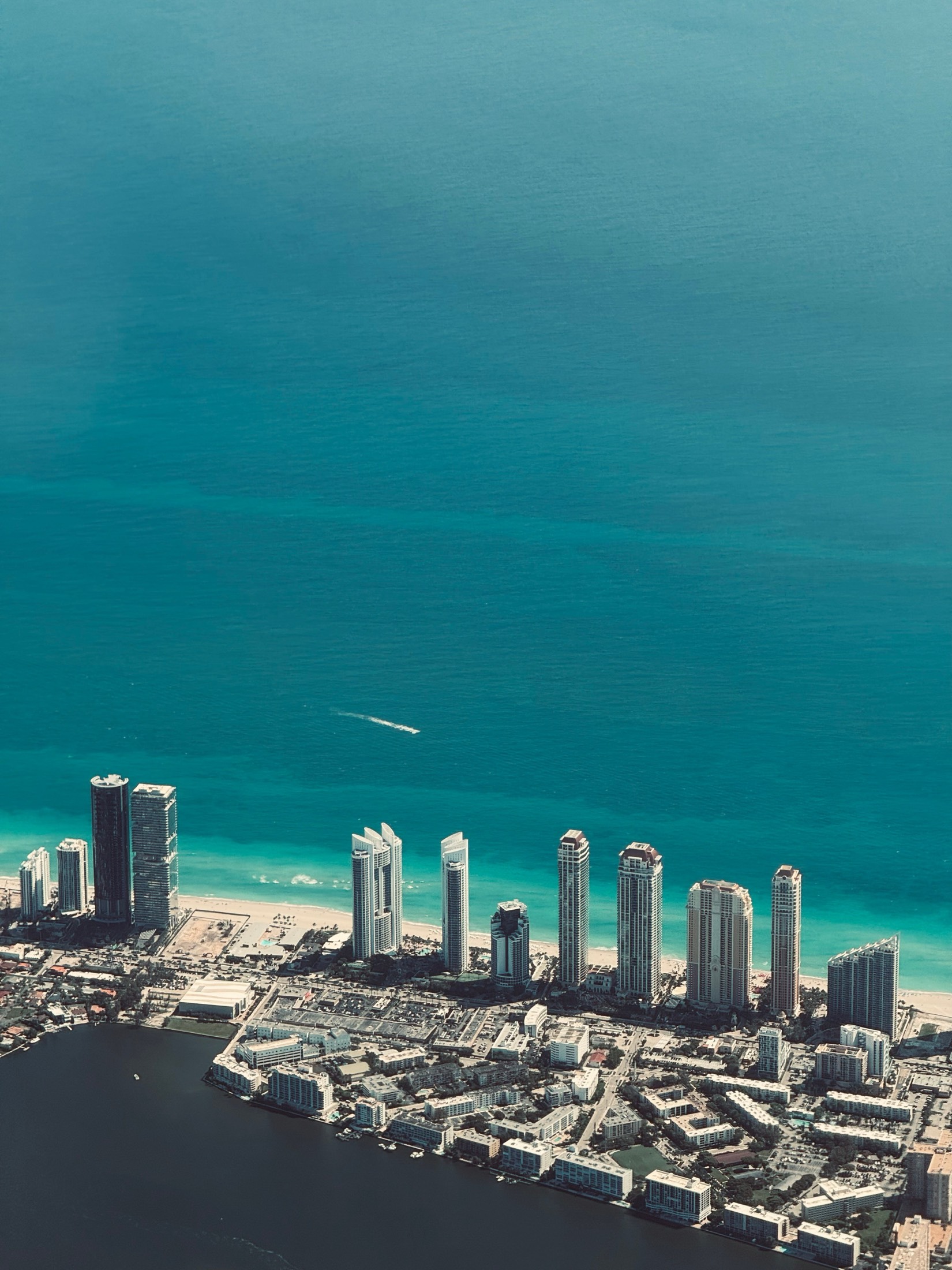 A line of beachfront towers and a marina at the edge of a turquoise sea, seen from the air over Miami