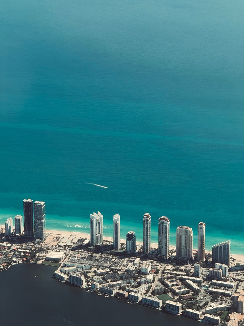 A line of beachfront towers and a marina at the edge of a turquoise sea, seen from the air over Miami