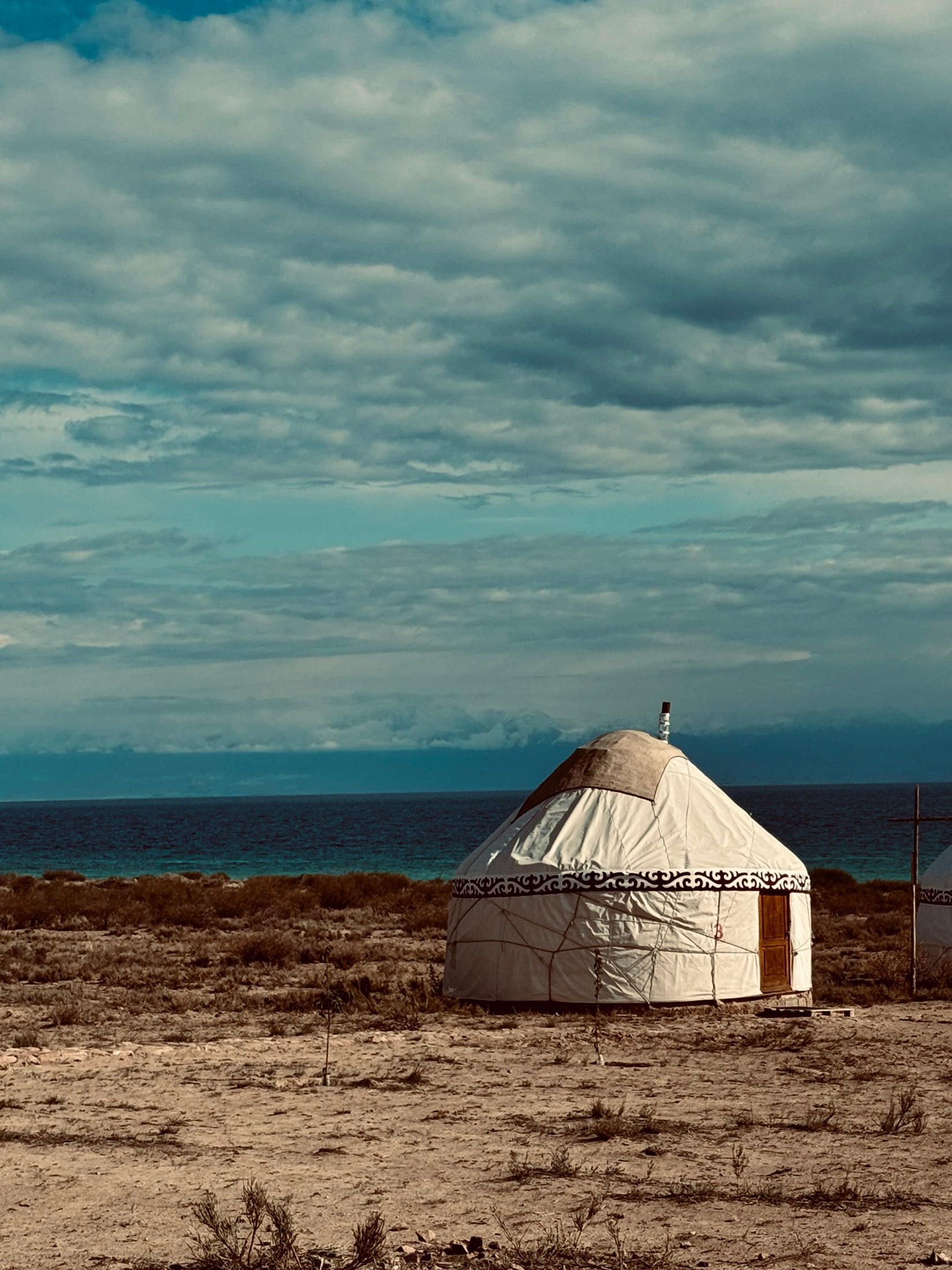 A white yurt on dry ground by the shore of a great lake under heavy clouds, Kyrgyzstan