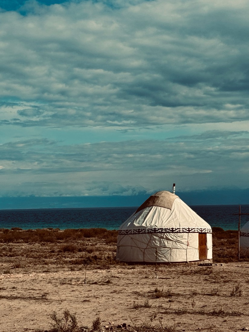 A white yurt on dry ground by the shore of a great lake under heavy clouds, Kyrgyzstan