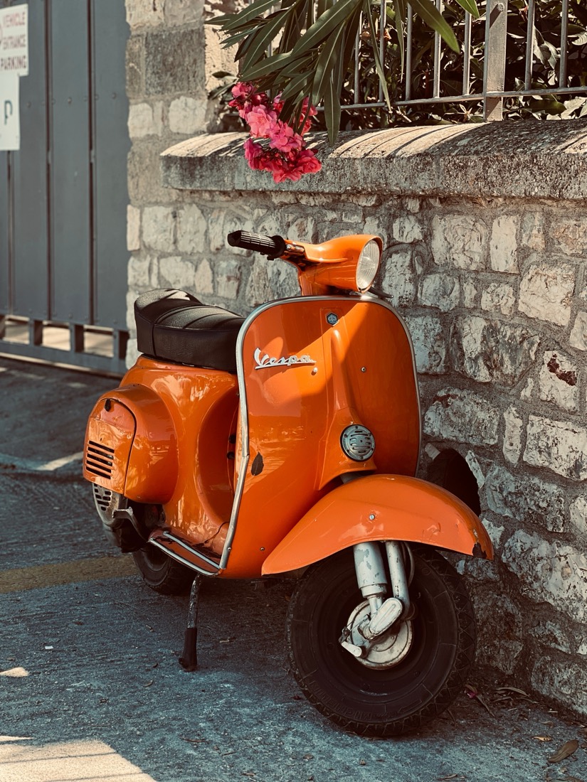 An orange Vespa parked against a stone wall under pink oleander on a Greek island street