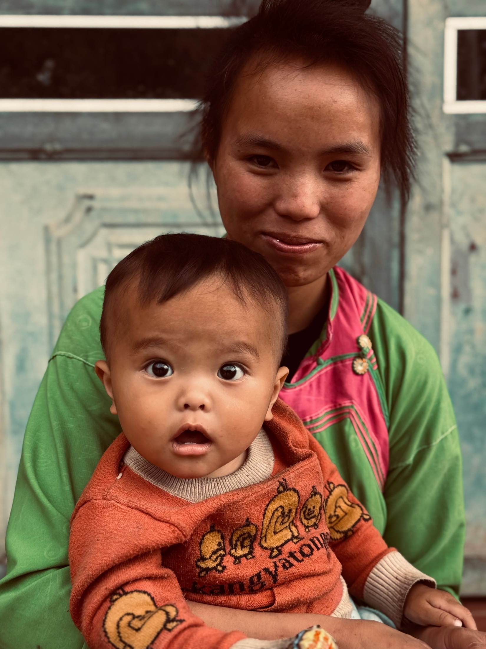 A Hmong mother holds her wide-eyed baby in an orange sweater in a doorway in the hills of northern Vietnam