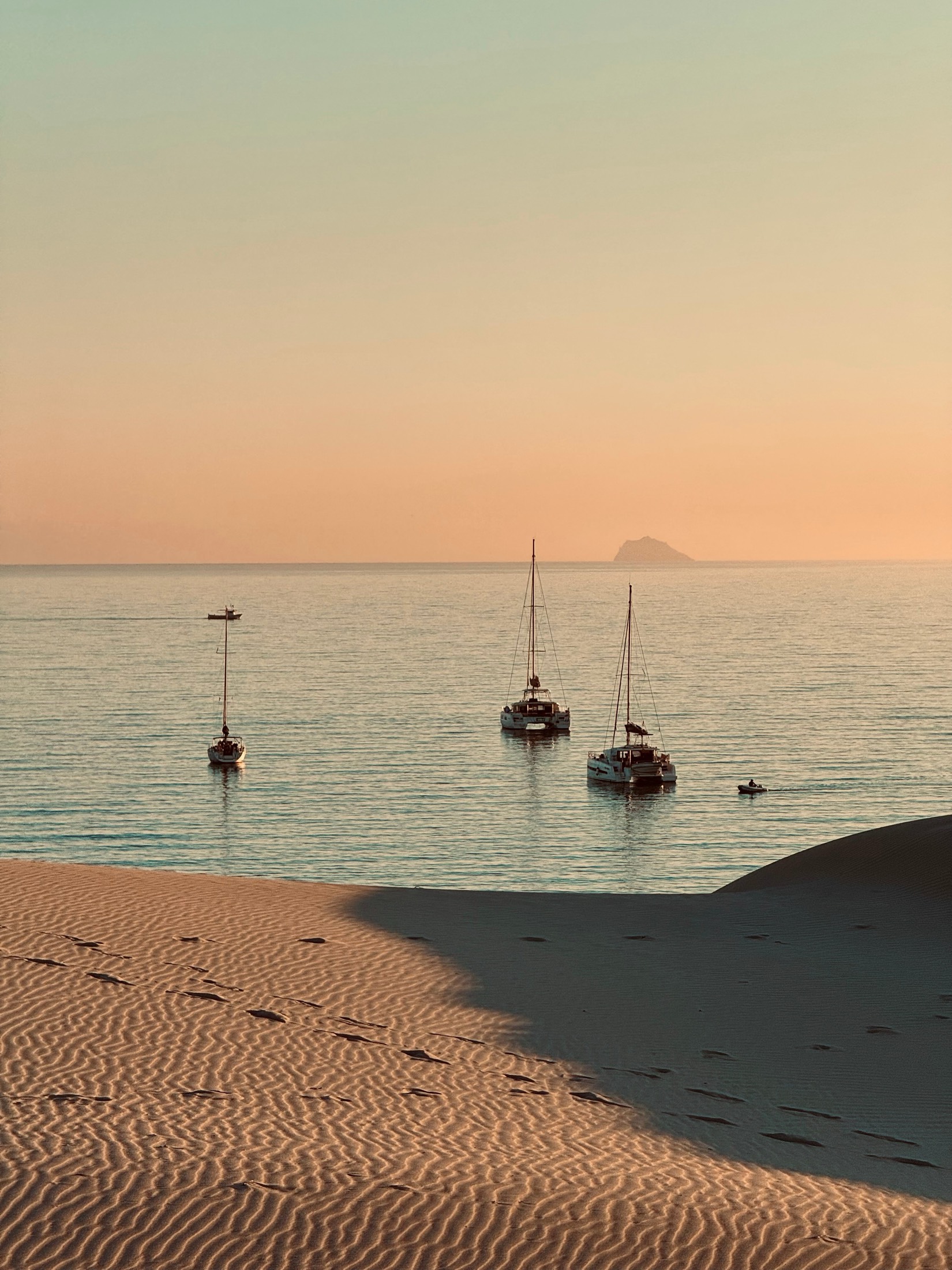 Footprints in a shadowed dune and three yachts at anchor on a pink evening sea, Sardinia