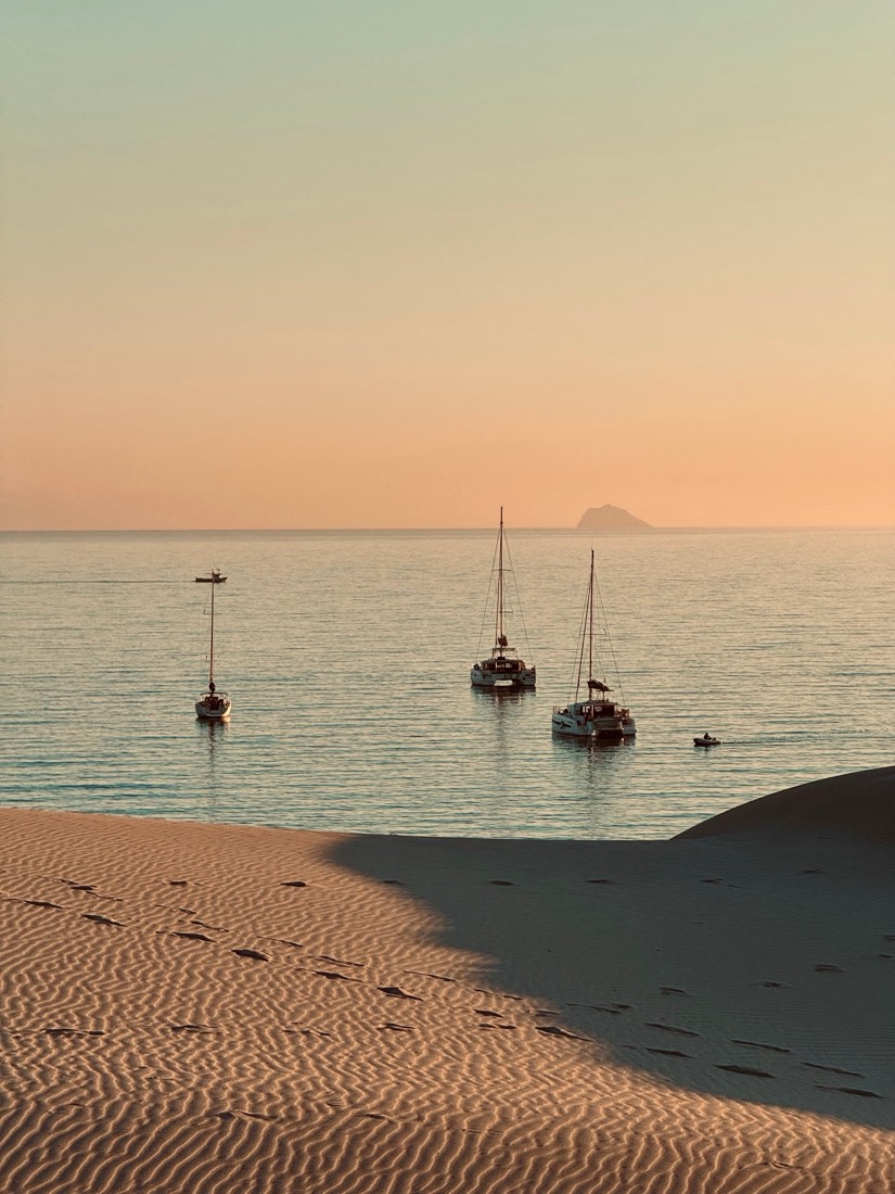 Footprints in a shadowed dune and three yachts at anchor on a pink evening sea, Sardinia