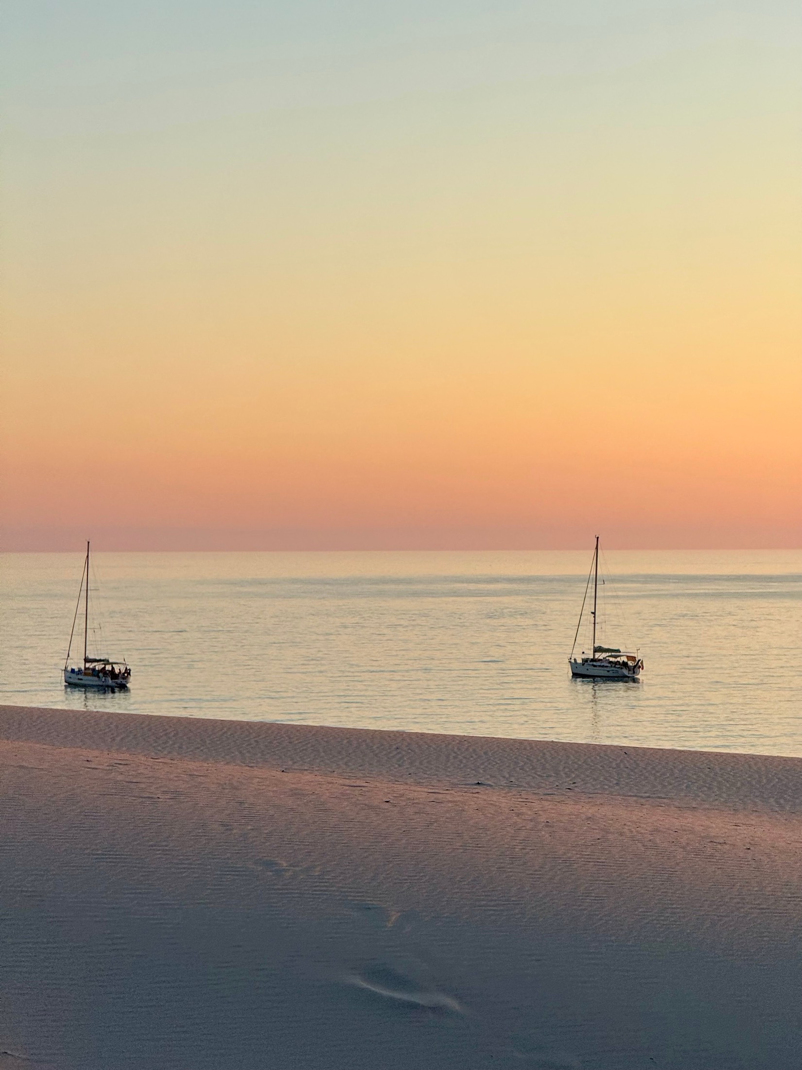 Two sailing boats on a calm sea at dusk seen from footprinted sand, Sardinia