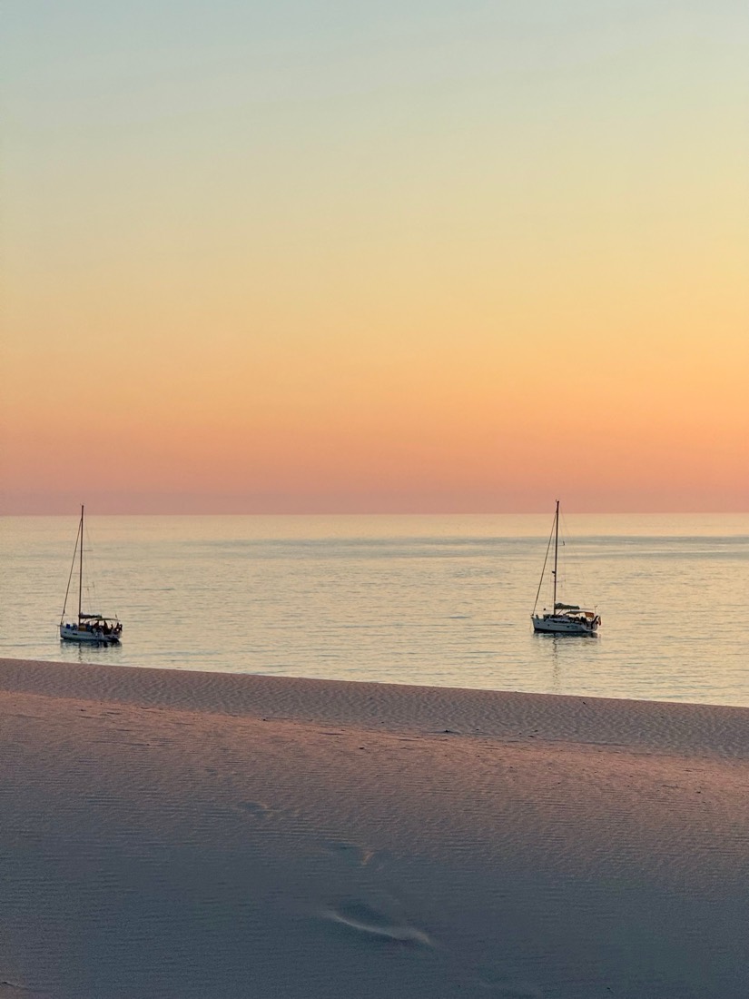 Two sailing boats on a calm sea at dusk seen from footprinted sand, Sardinia