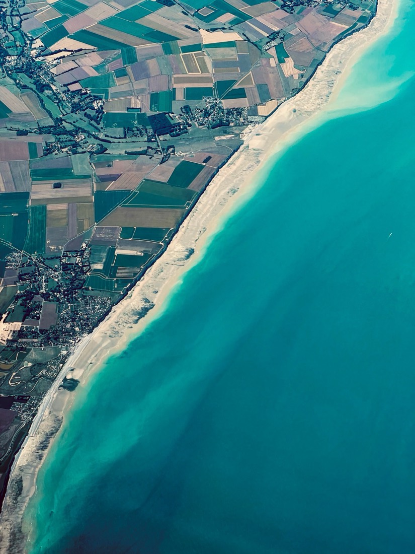A turquoise sea meets a long white beach and a patchwork of fields, seen from the air