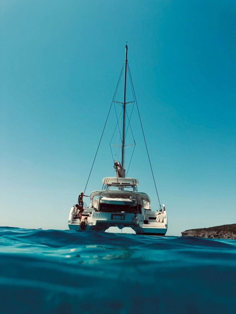 A sailing catamaran seen from water level, its bow lifting over a deep blue swell, Sardinia