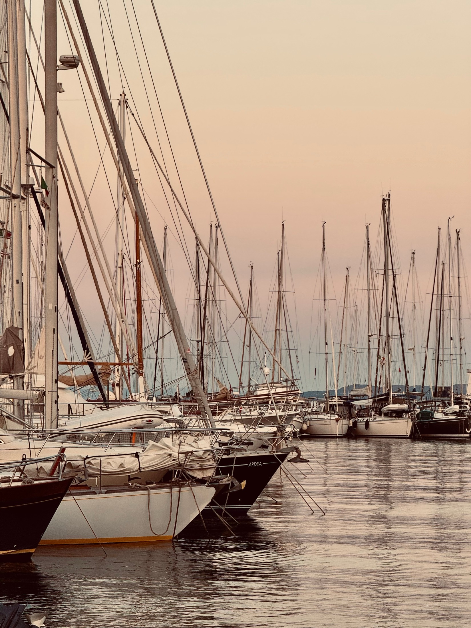 Masts and rigging of moored yachts against a soft pink evening sky in a Sardinian harbour