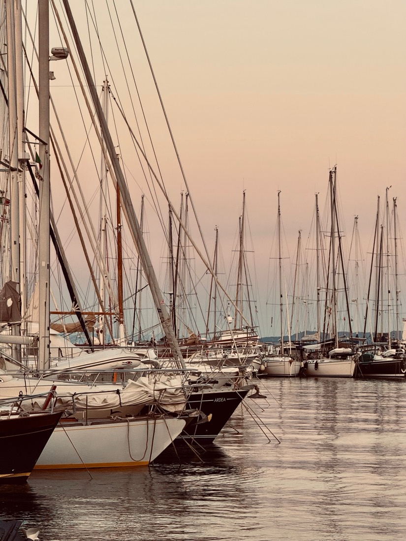Masts and rigging of moored yachts against a soft pink evening sky in a Sardinian harbour