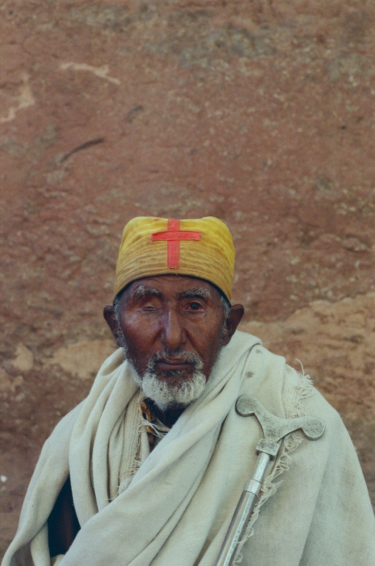 Elderly priest in a yellow cap with a red cross, wrapped in a white shawl, shot on film