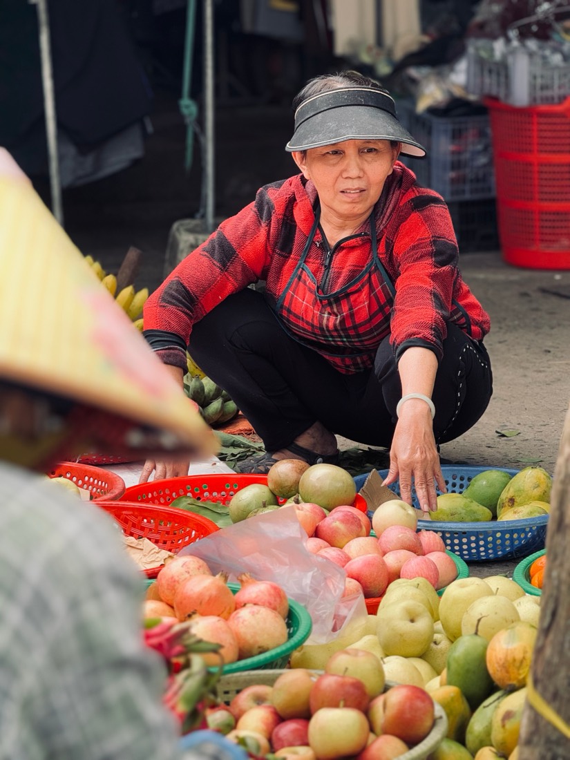 A woman in a red check shirt crouches over baskets of apples and fruit at a market in central Vietnam