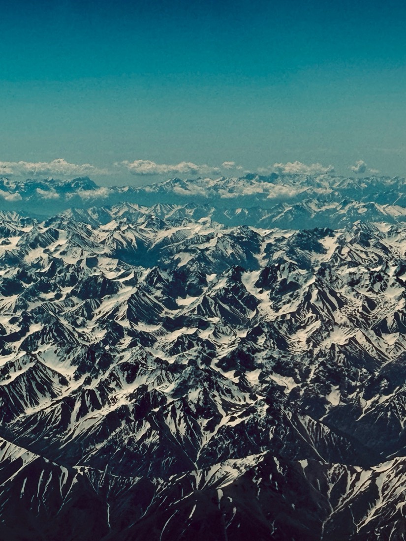 Snow-streaked mountain ranges rolling to the horizon under a deep blue sky, seen from a plane over northern Pakistan