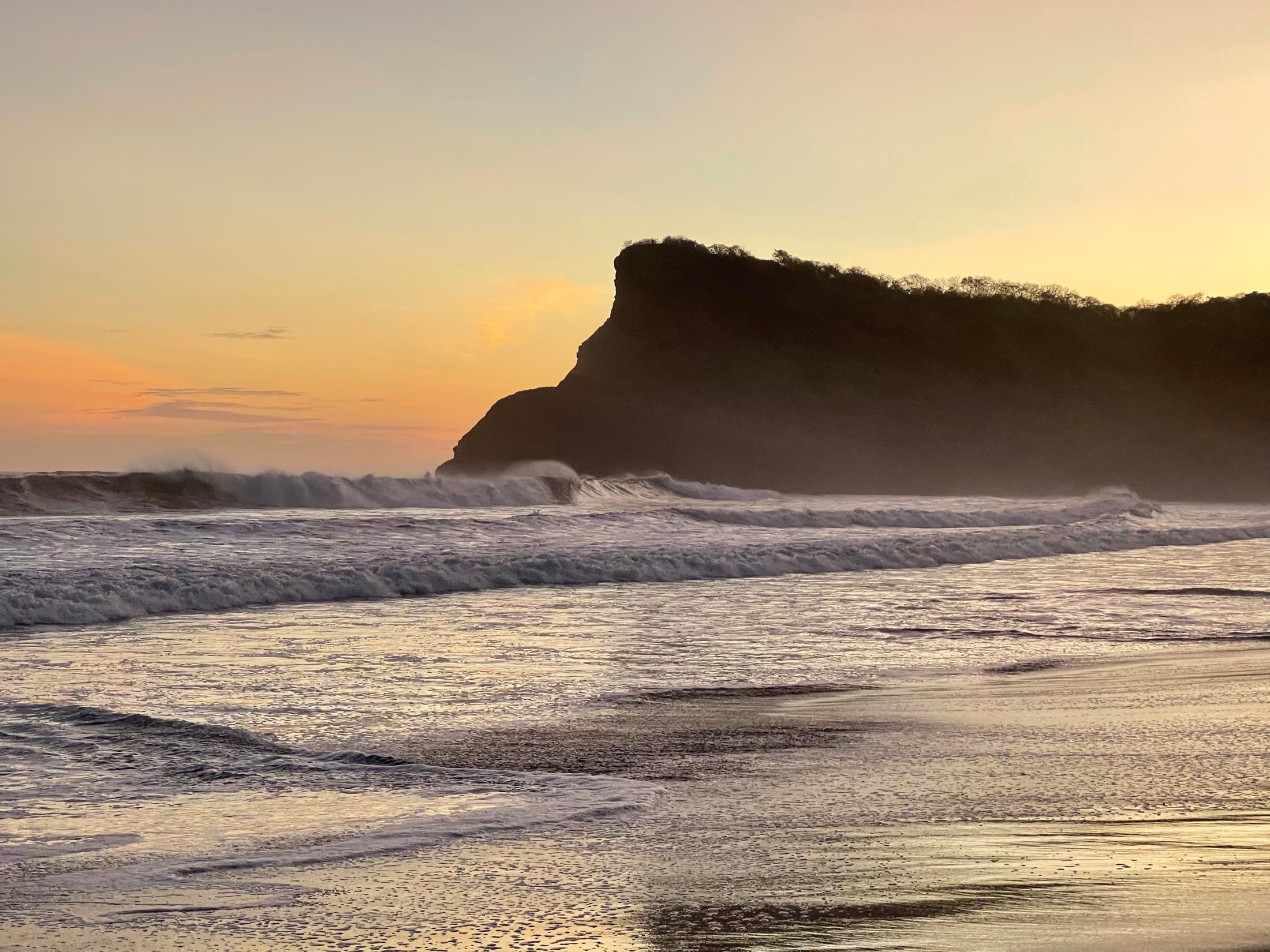 Surf rolls in toward a dark headland under a gold sunset sky on the Pacific coast of Nicaragua
