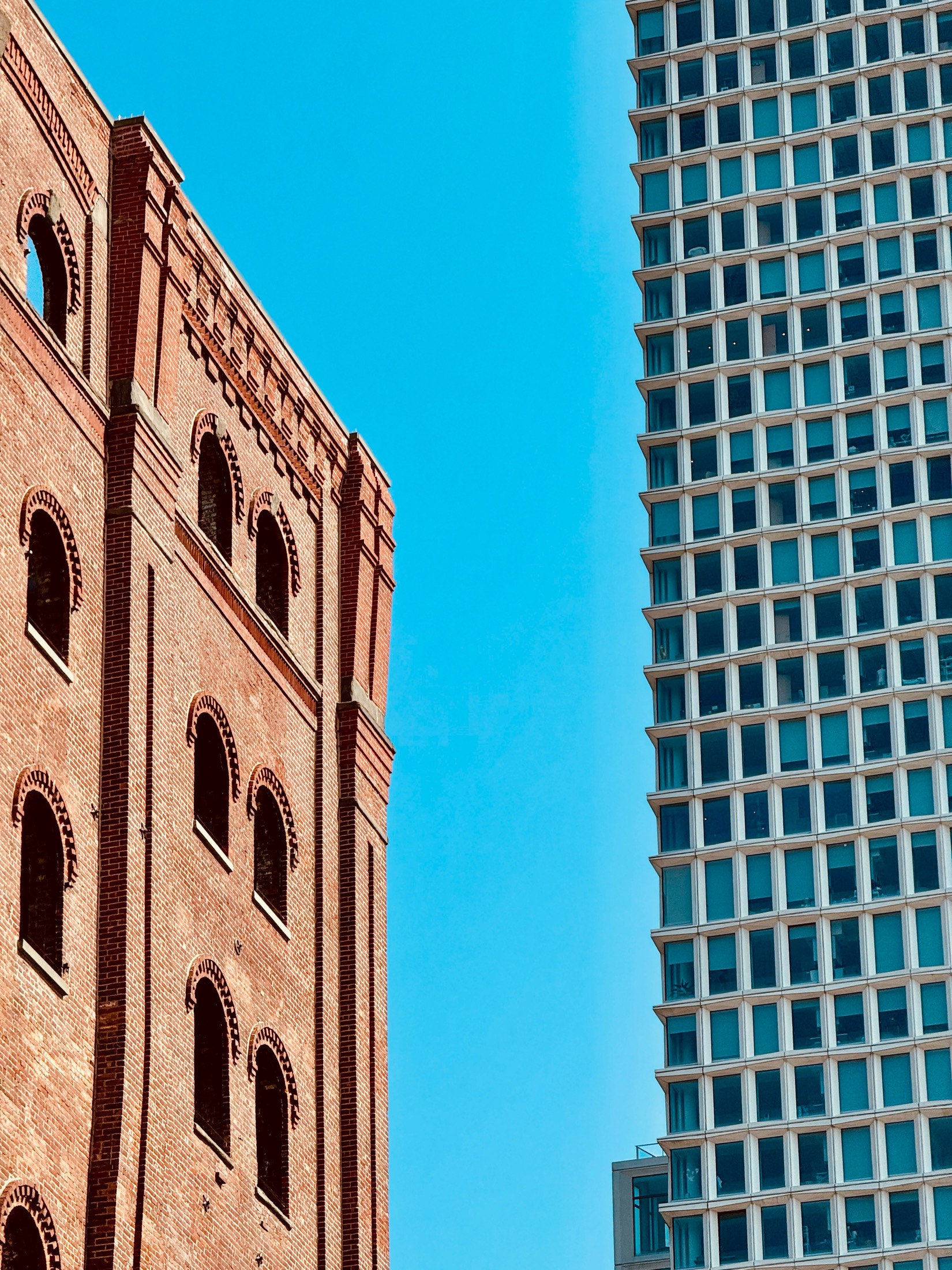 An old red-brick warehouse with arched windows beside a glass tower, against a blue sky in Brooklyn