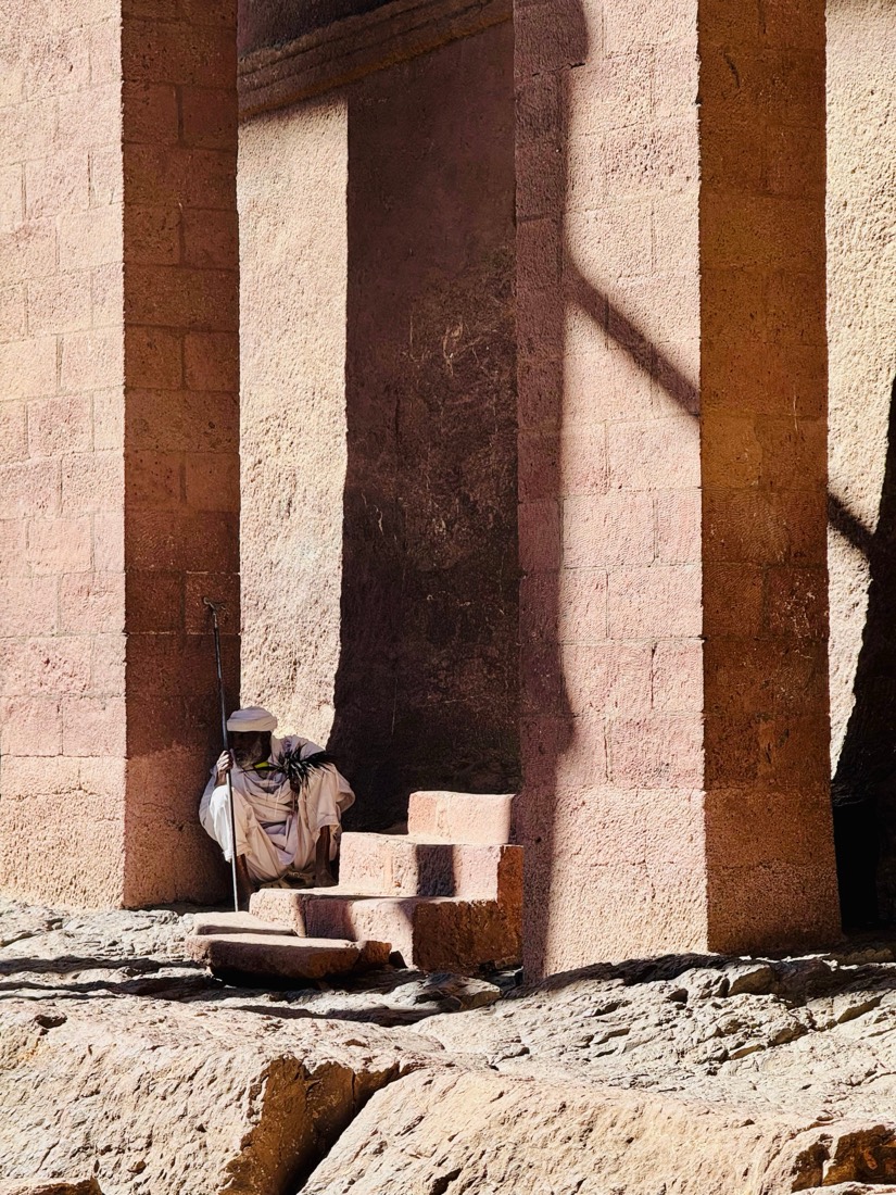 An elderly priest in white robes and turban crouches with his staff between towering pink stone pillars of a rock-hewn church, Lalibela, Ethiopia