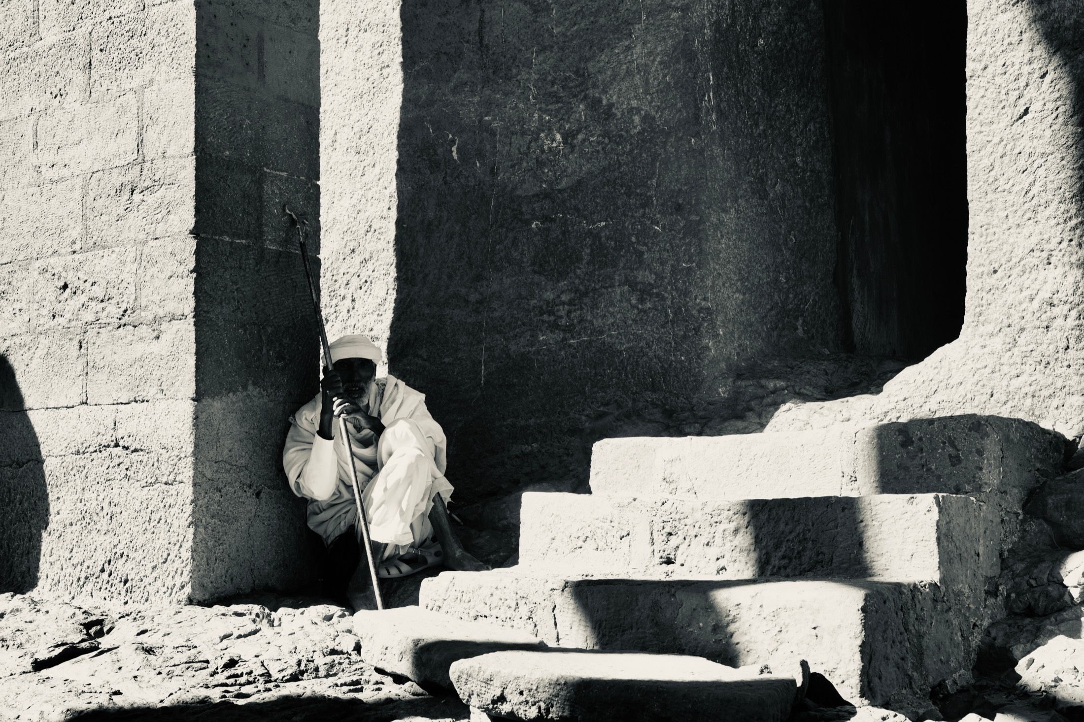 Black and white: an elderly priest in white robes rests on his staff beside worn stone steps in deep shadow at a rock-hewn church, Lalibela, Ethiopia