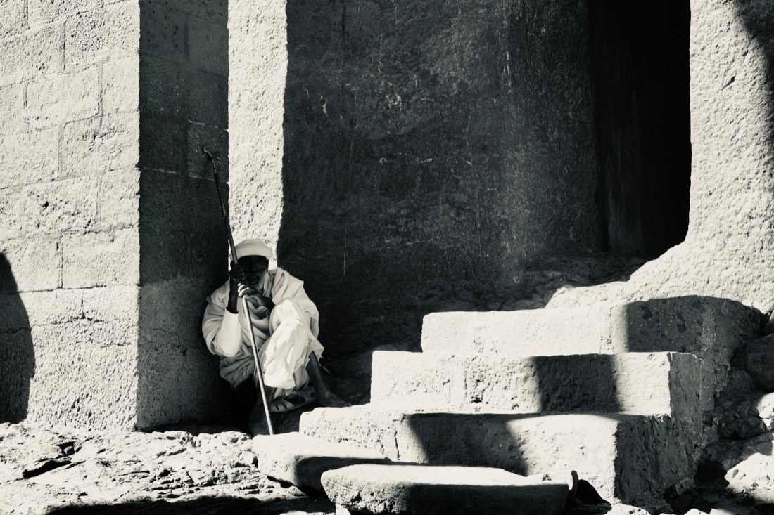 Black and white: an elderly priest in white robes rests on his staff beside worn stone steps in deep shadow at a rock-hewn church, Lalibela, Ethiopia