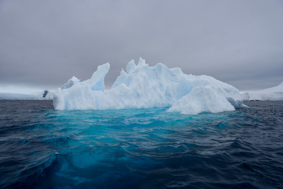 A jagged white iceberg under a grey sky, its submerged ice glowing turquoise beneath dark, choppy water, Antarctica