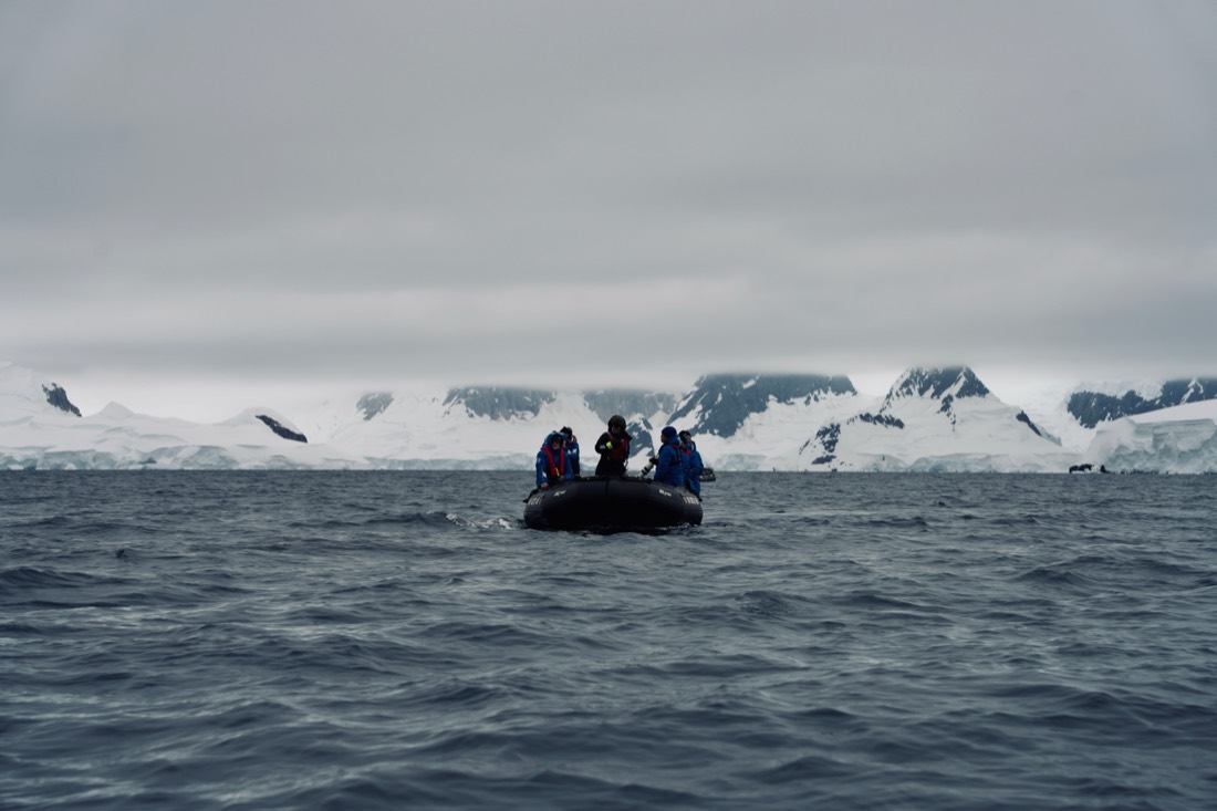 An inflatable Zodiac with a small expedition party in blue parkas crossing grey water towards snow-covered mountains, Antarctica