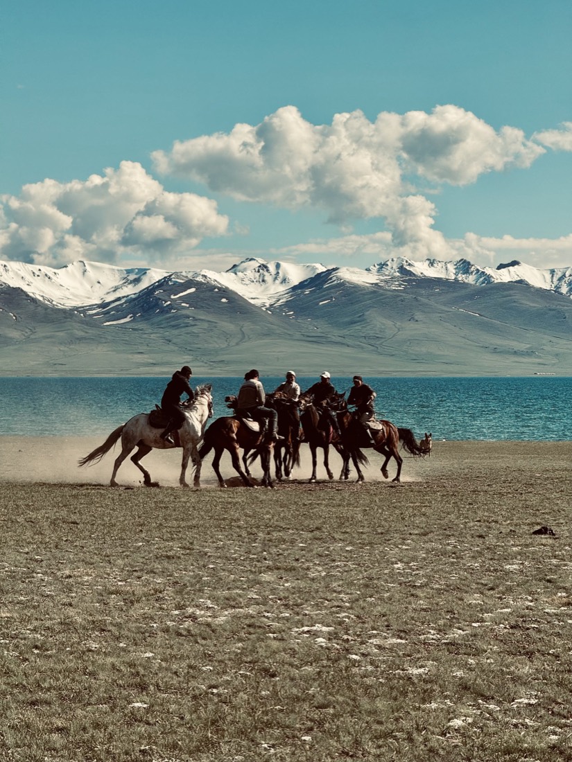 Horse riders crossing a beach below snow-capped mountains
