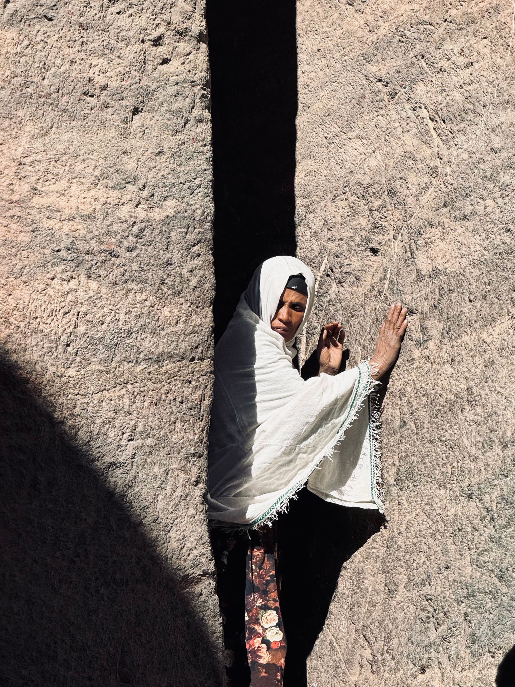 Woman wrapped in a white shawl pressed into a narrow passage of rock-hewn stone, hand raised to the wall