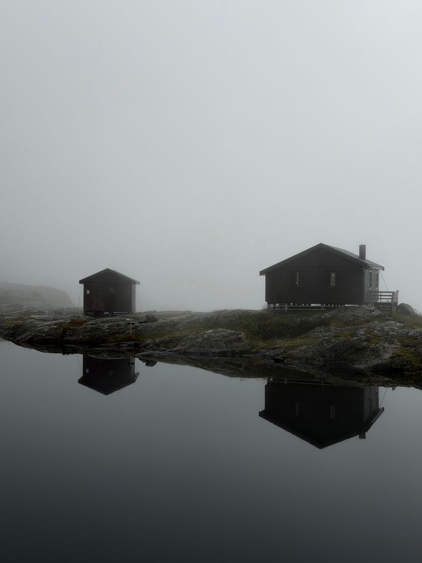 Two dark wooden cabins on rock above a mirror-still tarn, wrapped in fog