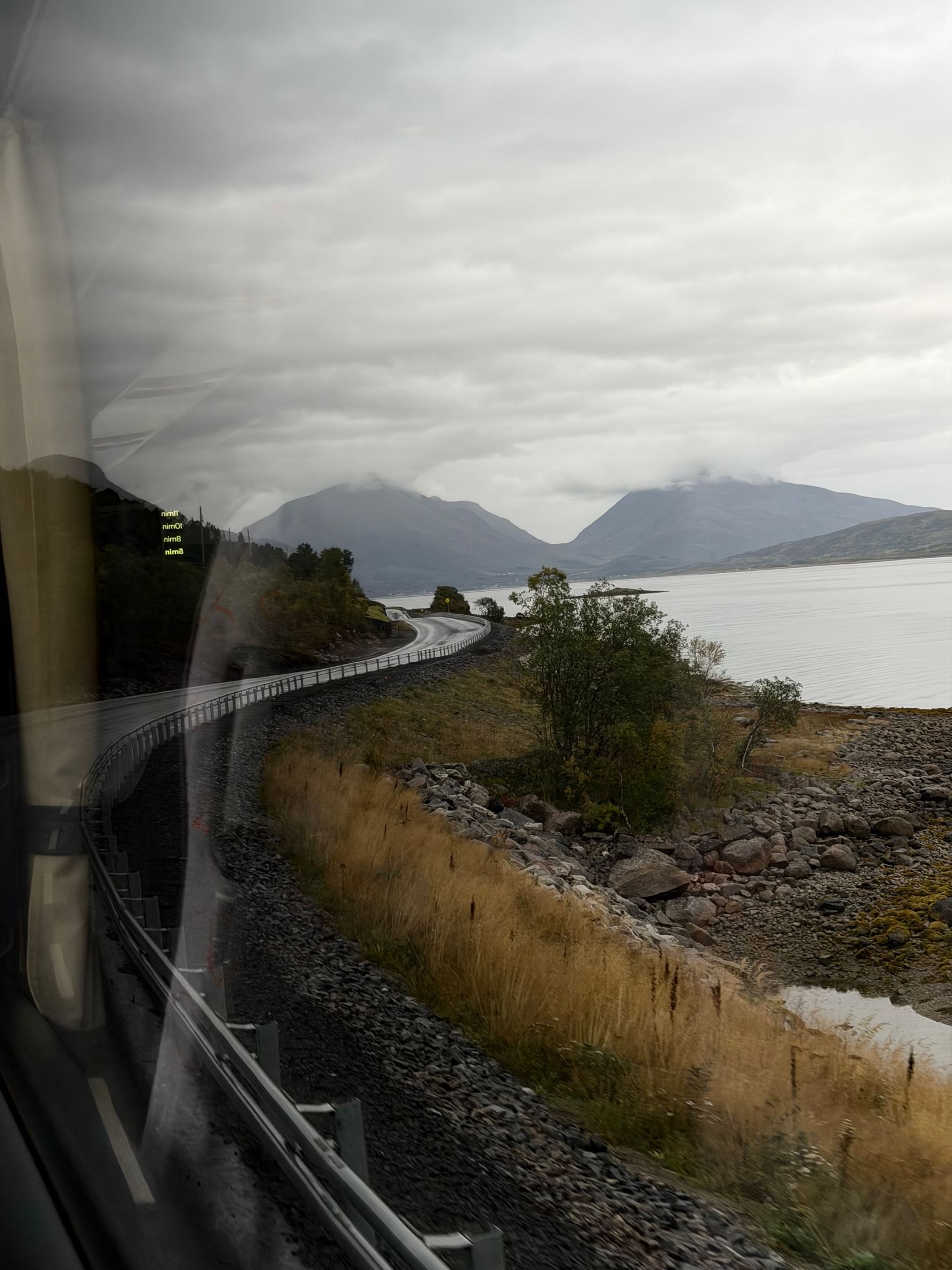 Lake, mountains and a curving rail line seen through a train window on a grey day, Lofoten, Norway