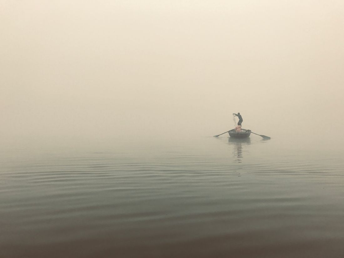 Lone fisherman standing in a small rowboat casting a net into thick fog over still water