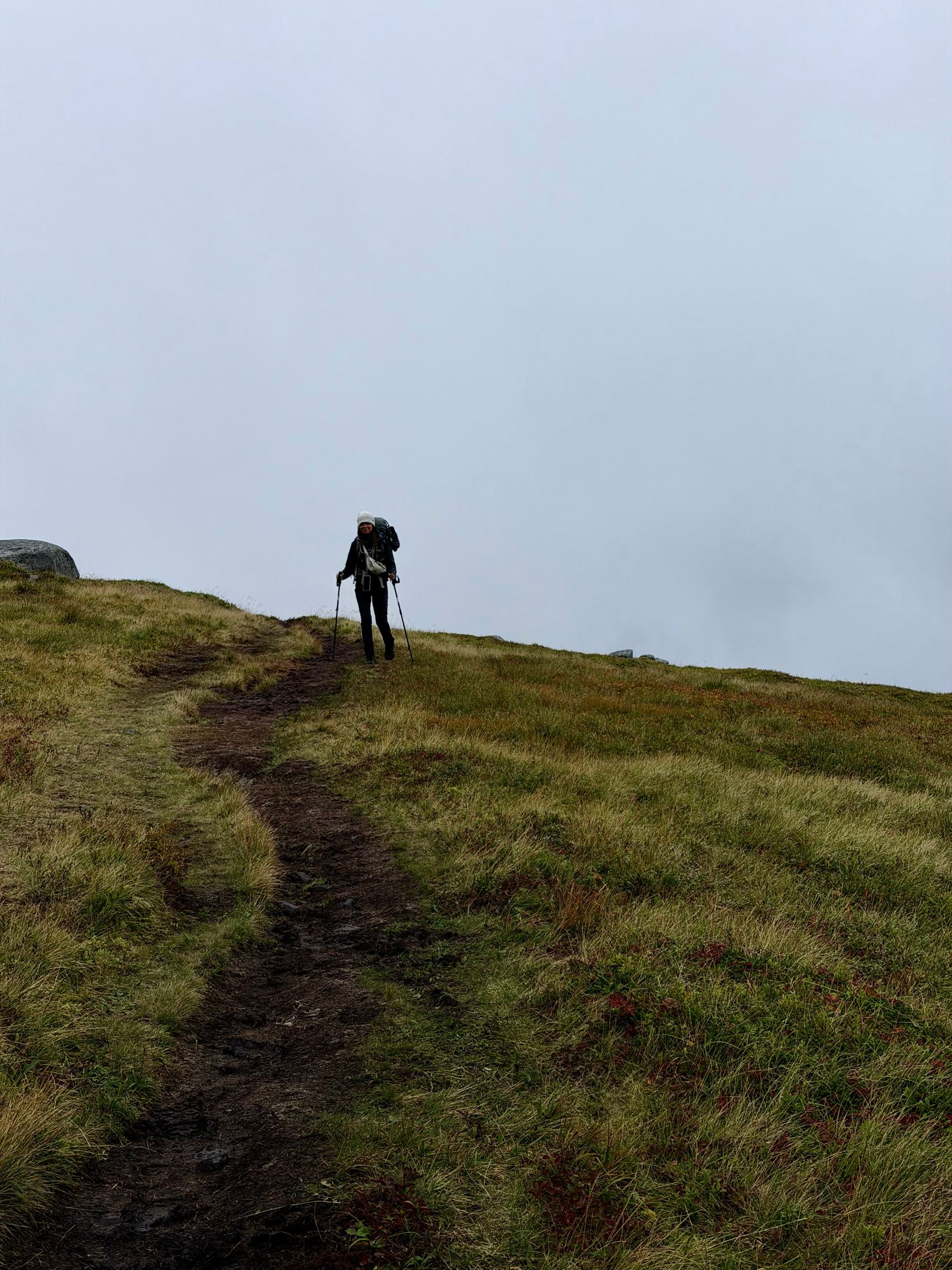 Lone hiker with trekking poles climbing a muddy grass trail into thick white fog, Lofoten, Norway