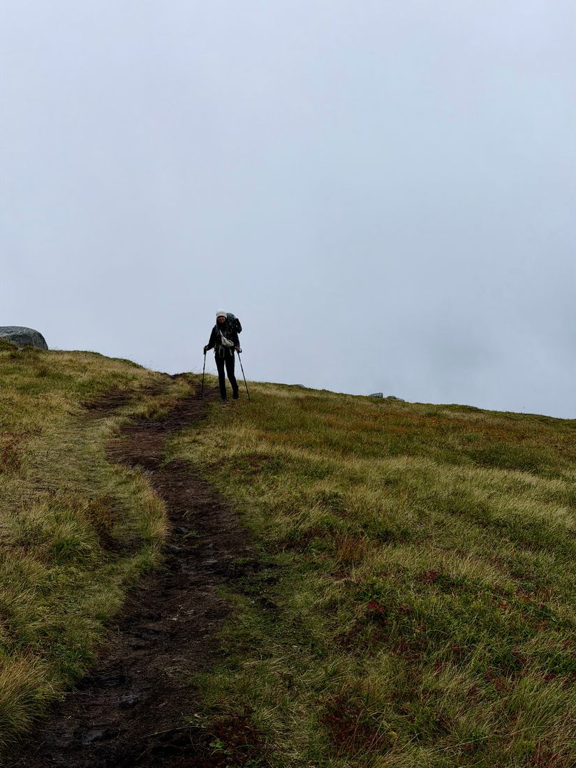 Lone hiker with trekking poles climbing a muddy grass trail into thick white fog, Lofoten, Norway