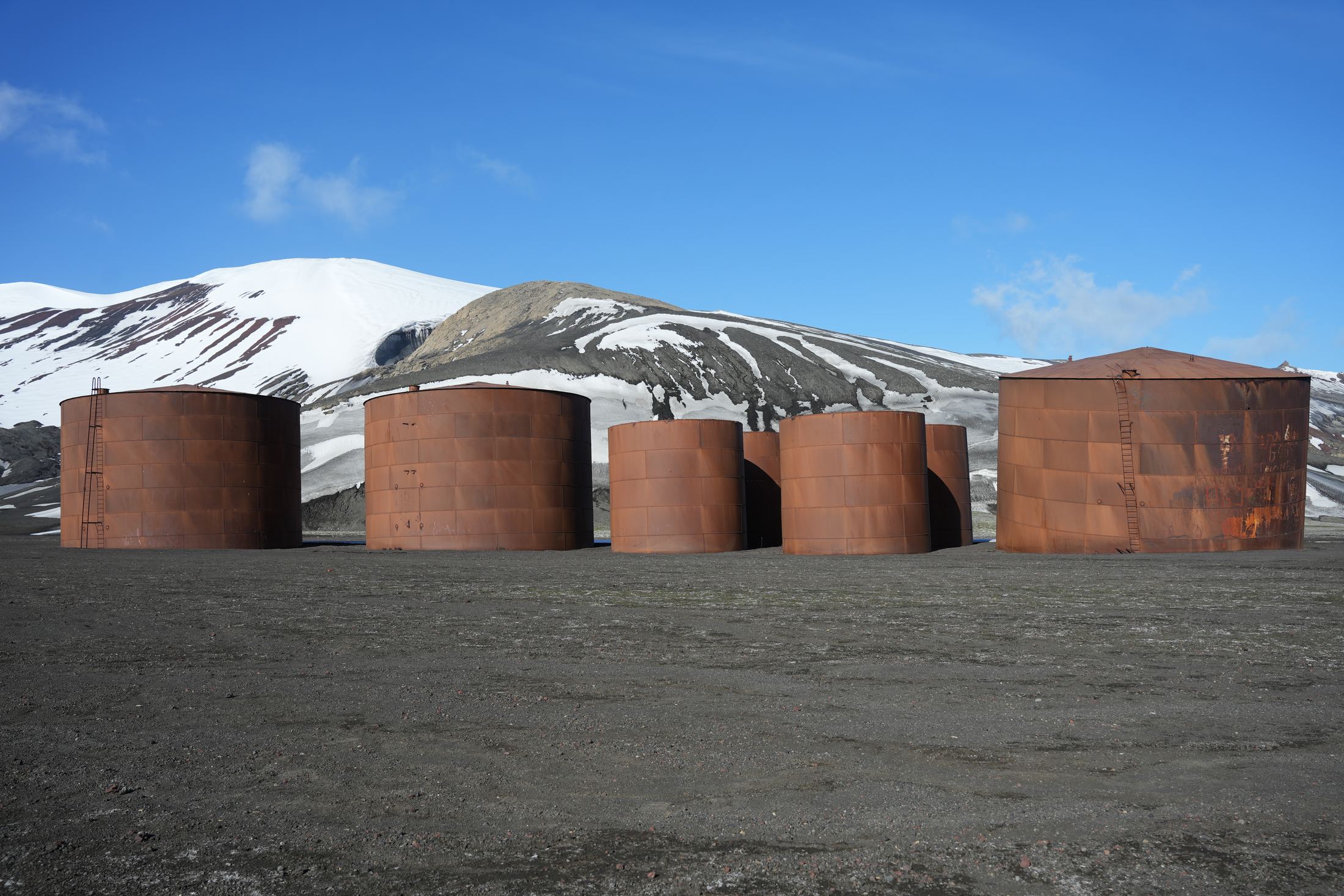 Row of rusted whaling-station tanks on a black volcanic beach below snowy hills