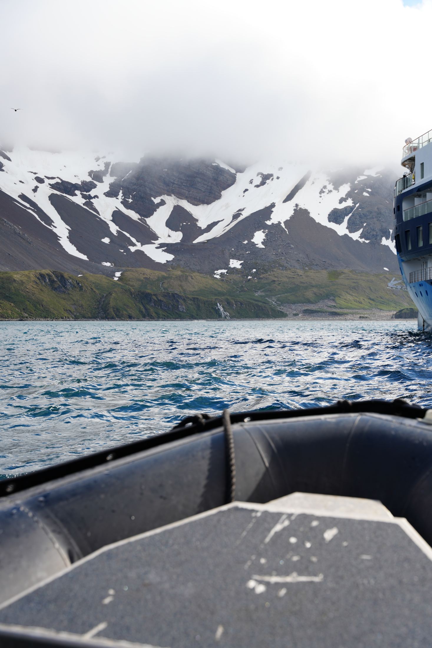 Bow of a rubber Zodiac pushing through choppy water toward snow-streaked mountains