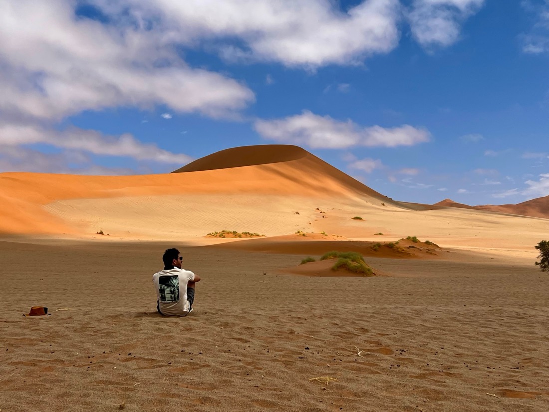A lone figure sits on pale sand facing a great orange dune under a blue sky, Namibia
